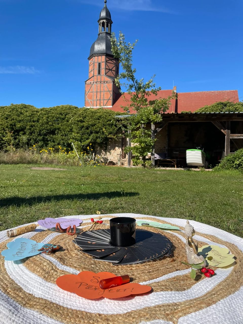 Mandala aus Karten, Kerze und Blüten auf einer Decke vor einem Backsteinkirchturm
